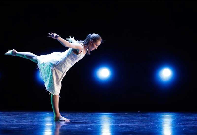 a student performing a dance piece leans out in front of a black background decorated with blue lights