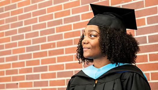 a woman in a graduation cap and gown stands in front of a brick wall